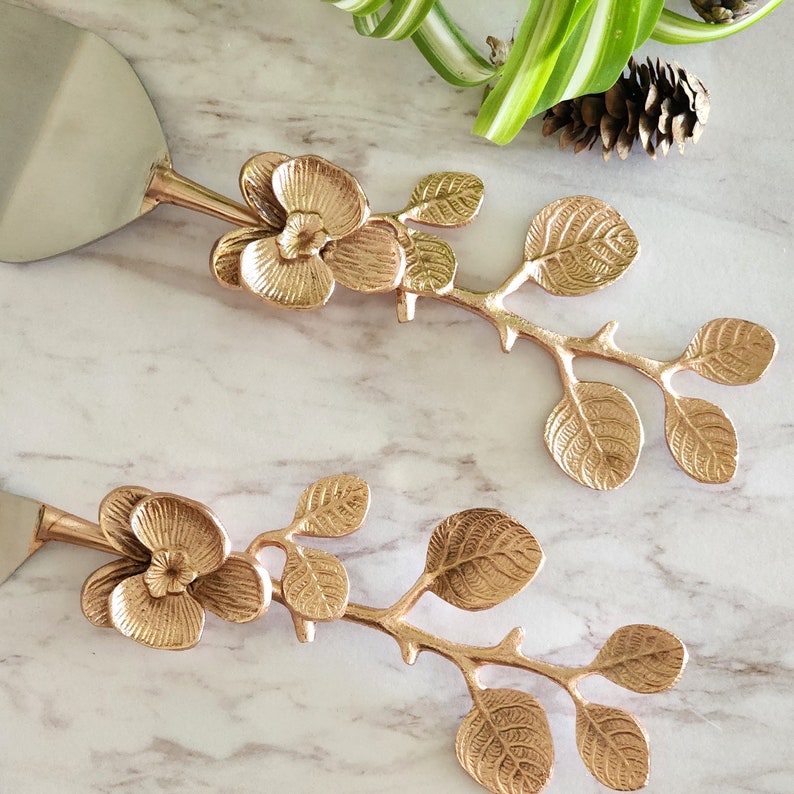 Puede incluir: Un juego de dos servidores de pastel de color dorado con dise&ntilde;os florales. Cada servidor presenta una flor delicada con hojas que se extienden desde el mango.