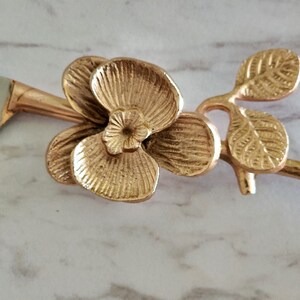 Puede incluir: Mango de cuchillo de metal dorado con dise&ntilde;o de flor de orqu&iacute;dea. El mango tiene forma de rama con dos hojas y una sola flor de orqu&iacute;dea.
