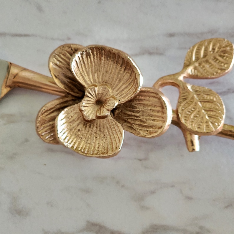 Puede incluir: Mango de cuchillo de metal dorado con dise&ntilde;o de flor de orqu&iacute;dea. El mango tiene forma de rama con dos hojas y una sola flor de orqu&iacute;dea.