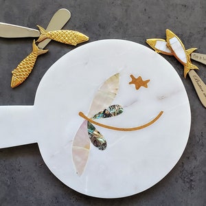 May include: White marble serving board with a gold dragonfly design and a gold star. The board has a handle and comes with three gold fish-shaped butter knives. The knives have the text "This is how we roll" and "Butter me up" engraved on them.