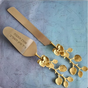 May include: A gold-colored cake server and knife set. The server has text in Hebrew. Both pieces have floral branch handles. The set is displayed on a blue map background. The server is approximately 10 inches long.
