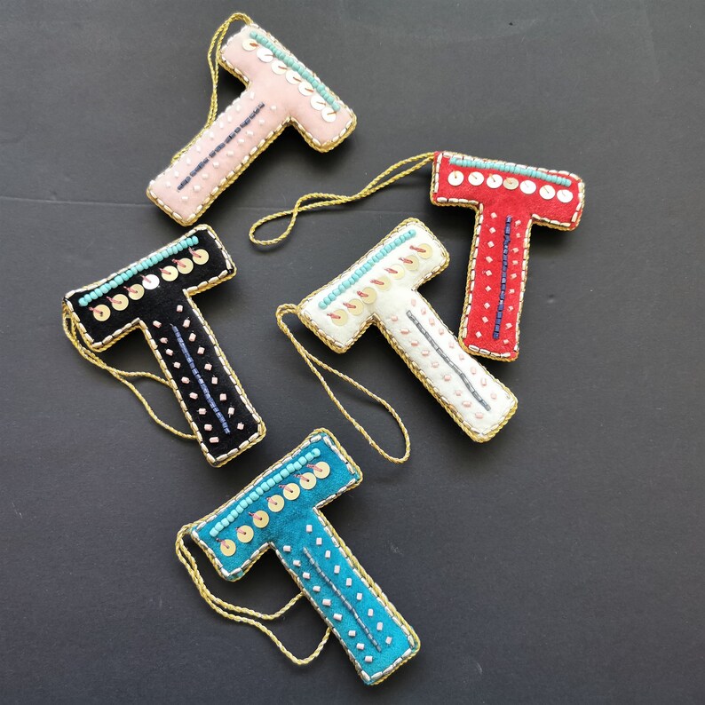 May include: Five felt ornaments shaped like the letter T, each decorated with different colored beads and sequins. The ornaments are pink, red, white, black, and teal.
