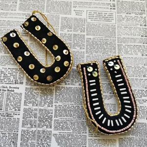 May include: Two black felt ornaments shaped like the letter U, decorated with gold and white beads and sequins. The ornaments are on a newspaper background.
