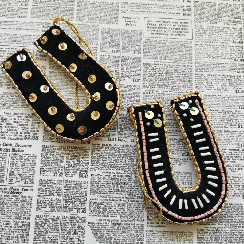 May include: Two black felt ornaments shaped like the letter U, decorated with gold and white beads and sequins. The ornaments are on a newspaper background.