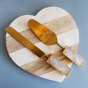 May include: A gold cake server and knife set with a white marble and wood heart-shaped serving board. The server has a floral design and the text "The Wrights" engraved on the handle. The handles are made of white quartz.