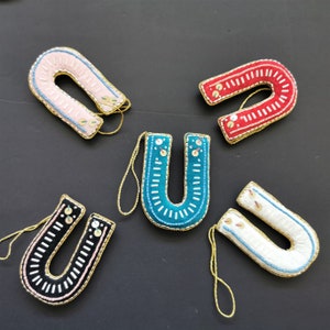 May include: Five felt horseshoe ornaments in different colors: pink, red, teal, white, and black. Each horseshoe is decorated with white and gold beads and has a gold string for hanging.