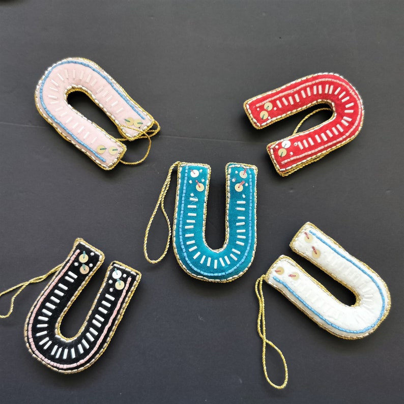 May include: Five felt horseshoe ornaments in different colors: pink, red, teal, white, and black. Each horseshoe is decorated with white and gold beads and has a gold string for hanging.