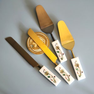 May include: Four cake servers with decorative handles. The handles are made of white marble with inlaid floral designs in gold, green, and blue. The blades are made of metal, two are gold and two are black.