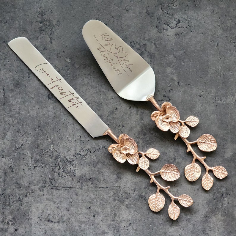 Puede incluir: Un juego de dos cuchillos para pastel de plata con mangos florales dorados. El cuchillo superior tiene el texto "Kiley & Nelson" y "2nd September 2023" grabado en &eacute;l. El cuchillo inferior tiene el texto "Love at first bite" grabado en &eacute;l.