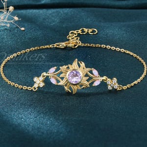 May include: Gold-toned bracelet featuring a sunburst design with a central purple gemstone. The bracelet includes small, delicate floral and leaf accents with clear and pink gemstones. The chain is fine and adjustable.