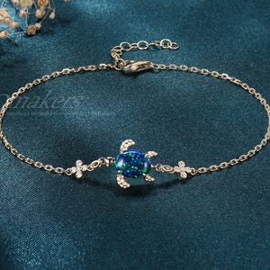 May include: A delicate gold-tone chain bracelet featuring a central sea turtle charm with a dark blue iridescent oval stone inlay. The turtle is flanked by two small four-petaled flower accents encrusted with tiny clear stones on each side.