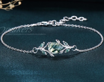 May include: A delicate silver bracelet featuring a central oval-shaped green gemstone, flanked by leaf-like accents. The bracelet has a fine chain and is set against a dark teal background, showcasing the jewelry's intricate design.