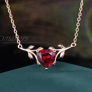 May include: A rose gold necklace featuring a heart-shaped, faceted red gemstone pendant. The pendant is framed by delicate leaf-like accents. The chain is a fine link style, and the necklace is displayed against a dark background.