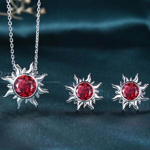 May include: A silver-toned jewelry set featuring a sunburst design with a large, round, red gemstone center. The set includes a necklace and a pair of matching stud earrings. The background is a dark teal fabric.