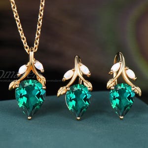 May include: A gold-toned jewelry set featuring a necklace and two earrings. Each piece has a teardrop-shaped, emerald-green gemstone, accented with white stones and leaf-like details. The set is displayed on a teal surface.