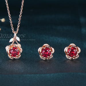 May include: A rose gold jewelry set featuring a necklace with a pendant and two matching stud earrings. The pendant and earrings have a rose design with a vibrant red center stone. The necklace chain is delicate.