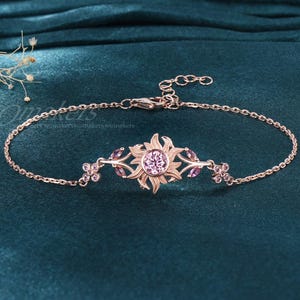 May include: A rose gold bracelet featuring a sunburst design with a central pink gemstone. The bracelet has delicate chain links and is adorned with small purple gemstones and floral accents. The jewelry is displayed on a teal fabric.