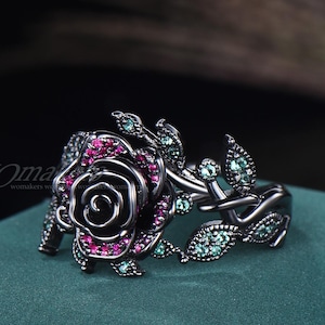 Könnte beinhalten: Schwarzer Ring mit Rosenmotiv, besetzt mit rosa und grünen Edelsteinen.