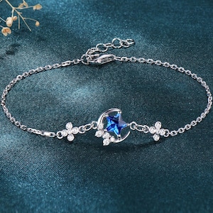 May include: A silver bracelet with a delicate chain and a central charm. The charm features a blue star within a crescent moon, surrounded by small, sparkling accents. Two flower-shaped embellishments with clear stones are on either side of the central charm.