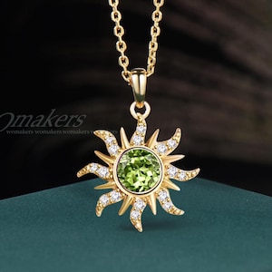May include: Gold-toned necklace featuring a sun pendant with a central green gemstone and surrounding white crystals. The sunburst design is detailed with pointed rays. The chain is delicate and the pendant is suspended from a small bail.