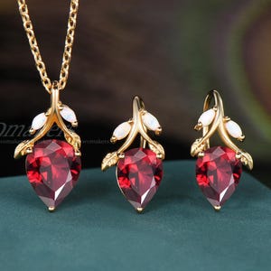May include: A gold-toned jewelry set featuring a necklace and two earrings. Each piece has a teardrop-shaped, deep red gemstone, accented with white stones and leaf-like details. The set is displayed on a teal surface.