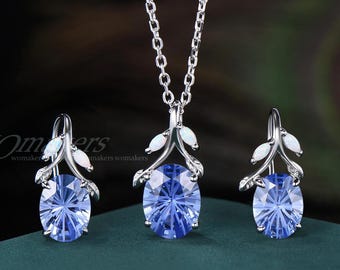 Könnte beinhalten: Ein Schmuckset bestehend aus einer silberfarbenen Halskette und passenden Ohrringen. Jedes Teil zeigt einen ovalen, facettierten blauen Stein in einer zweigartigen Fassung mit kleinen weißen Akzenten. Der Text lautet WOmakers.