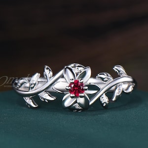 May include: A silver-toned ring featuring a central flower motif with a small, round, vibrant red gemstone at its core. The band is designed with sculpted, overlapping leaf patterns that extend outward from the flower on both sides of the ring.