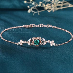 May include: A delicate rose gold bracelet with a green gemstone surrounded by white gemstones. The bracelet features a floral design with leaves and a chain link.