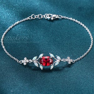 May include: A silver bracelet featuring a central oval-cut, deep red gemstone. The bracelet has a delicate chain design with small floral and leaf accents, and a secure clasp. The background is a teal-colored fabric.