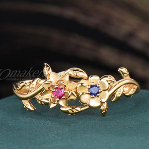 May include: A gold-colored ring featuring a floral design with two flowers. One flower has a pink gemstone center, and the other has a blue gemstone center. The ring has a delicate, nature-inspired aesthetic.
