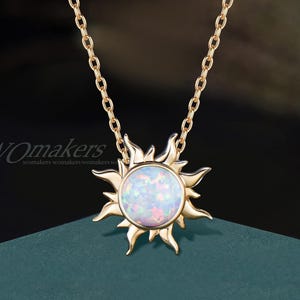 May include: A gold-colored necklace featuring a sun pendant with a central opal stone. The sunburst design has pointed rays around the opal. The chain is also gold-colored. The brand name "WOMakers" is visible in the lower left corner.