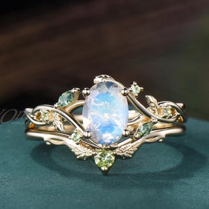 May include: A gold ring featuring a central oval-cut iridescent moonstone, flanked by small green peridot gemstones and marquise-cut moss agate stones. The band is designed with intricate metal leaf and vine motifs wrapping around the stones.