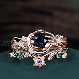 May include: A rose gold ring set featuring a dark blue star-shaped stone, a crescent moon, and small white opals. The band includes intricate vine-like metalwork with green stones and a small purple stone at the base. The text WOmakers is visible.