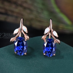 May include: A pair of rose gold earrings with a leaf design. Each earring features a large, oval, deep blue gemstone and two small white gemstones. The earrings are displayed on a dark green surface.
