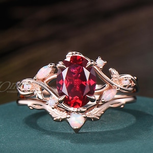 May include: A rose gold ring set featuring a central oval-cut deep red gemstone held by prongs. The band is designed with leaf-like metalwork and accented with small, iridescent oval-shaped opals arranged along the sides and the base of the setting.