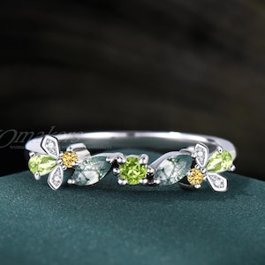 May include: A silver ring featuring a floral design with green and yellow gemstones. The ring has a delicate band and is adorned with leaf-shaped and round gemstones. The ring is set against a dark green background.
