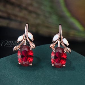 May include: A pair of rose gold earrings featuring oval-cut, deep red gemstones. Each earring has a leaf-like design with white accents. The earrings are displayed on a dark green surface, with a blurred background.