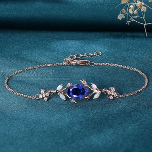 May include: A delicate rose gold bracelet with a blue oval gemstone surrounded by white opal accents. The bracelet features a floral design with small leaves and a chain link.