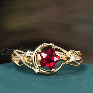 May include: A gold ring featuring a central star-shaped red gemstone set within a crescent moon frame. The band is crafted with intricate, swirling metalwork and accented with three small, round, iridescent white stones along the sides.