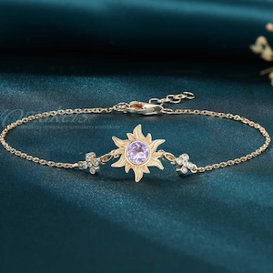 May include: Gold-toned bracelet featuring a sun-shaped charm with a purple center stone. The bracelet has delicate chain links and small floral accents on either side of the sun. The clasp is adjustable.