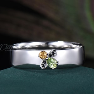 May include: A silver ring with a flat band, featuring a bee design. The bee has a yellow gemstone head, a green pear-shaped gemstone body, and black gemstone wings. The ring is set against a dark green surface.