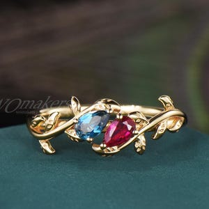 May include: A gold-colored ring featuring a vine design with two pear-shaped gemstones. One gemstone is a vibrant blue, and the other is a deep red. The ring has a delicate, nature-inspired aesthetic.