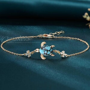 May include: Gold-toned bracelet featuring a turquoise turtle charm with black accents. The bracelet includes small, clear gemstone accents and a delicate chain. The clasp is adjustable. The bracelet is displayed on a teal background.
