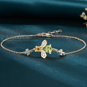 May include: Gold-toned bracelet featuring a bee-shaped pendant with a green gemstone body, yellow accents, and clear stone wings. The bracelet includes delicate chain links and small floral accents on either side of the bee.