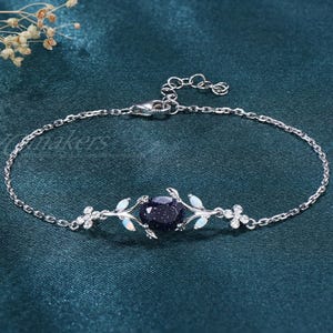 May include: A silver bracelet with a dark blue gemstone center. The bracelet features delicate floral and leaf accents with small white stones. The adjustable chain is displayed on a teal-colored fabric.
