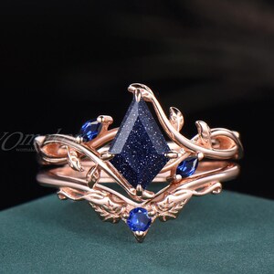 May include: Rose gold ring set featuring a large, dark blue gemstone with a sparkling effect, set in a diamond shape. The ring is adorned with smaller blue gemstones and delicate leaf-like details.