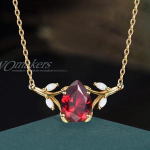 May include: A gold chain necklace with a red gemstone pendant. The pendant is shaped like a teardrop and is surrounded by small white gemstones. The pendant is attached to a delicate gold branch design.