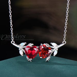 May include: A silver necklace featuring two heart-shaped, faceted red gemstones. The hearts are connected by a silver branch design with small white accents. The necklace hangs on a delicate silver chain.