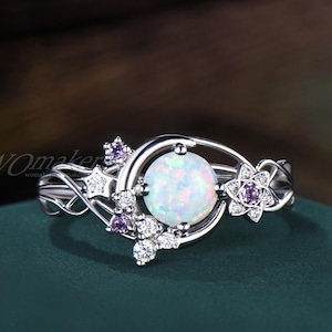 May include: A silver-toned ring featuring a central round iridescent opal stone, accented by a crescent moon shape, small purple round-cut stones, and clear star-shaped and round-cut stones set along a delicate, intertwining metal band.