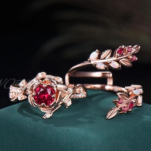 May include: Rose gold rings with floral and leaf designs. One ring features a large, round, red gemstone at the center of a rose. Other rings have leaf and gemstone accents. The rings are set on a dark green surface.